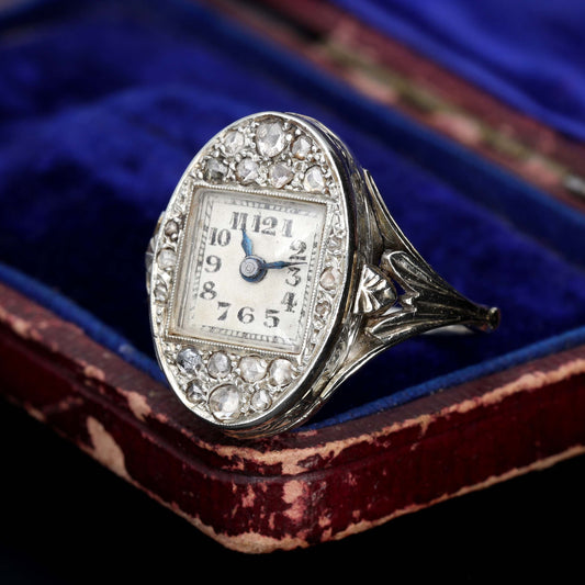 Art Deco diamond ring watch with oval bezel, rose-cut diamond halo surrounding a square cream dial with Arabic numerals and blued steel hands