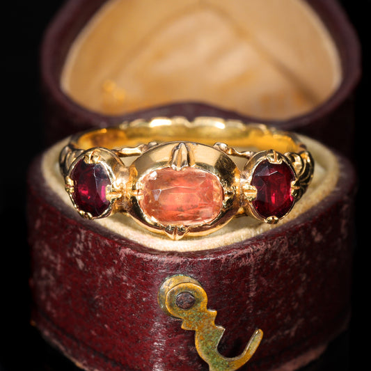 Antique French 19th-century three-stone ring with a central pinkish-peach topaz flanked by two deep red garnets in yellow gold, displayed in a period burgundy ring box